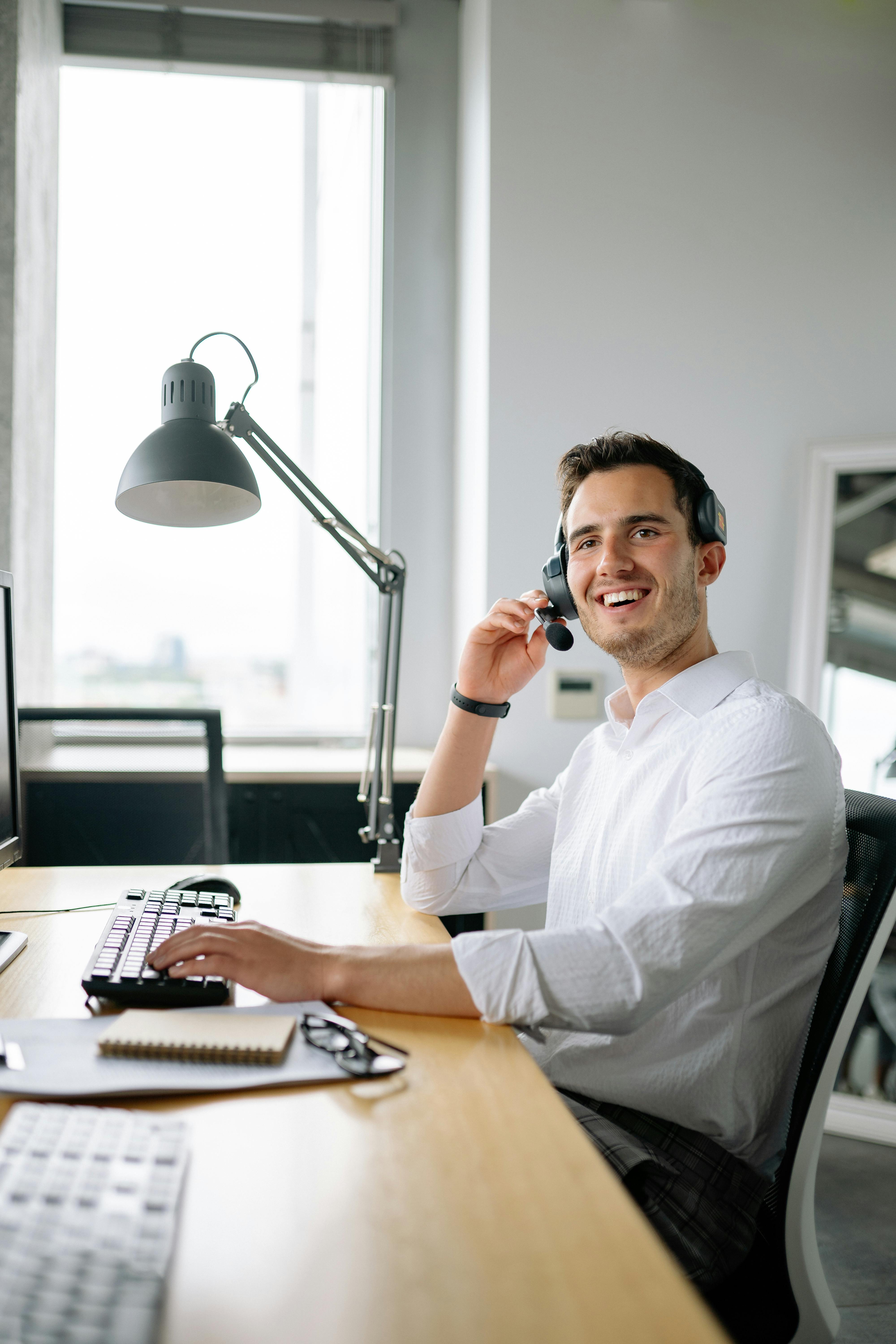 A cheerful call center agent wearing headphones works at a modern office desk.